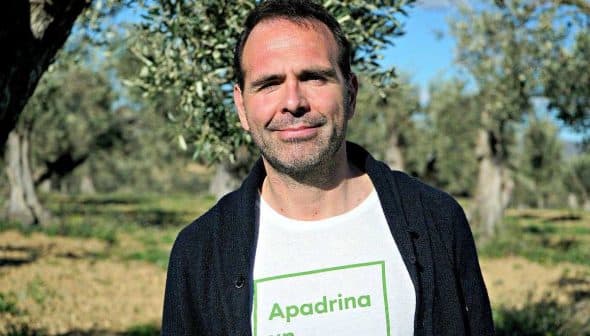 Man standing in an olive grove wearing a white t-shirt with green text. - Olive Oil Times