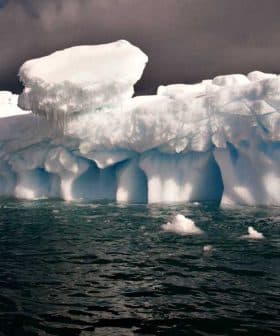 A large iceberg floating in ocean water with a blue hue and cloudy sky above. - Olive Oil Times