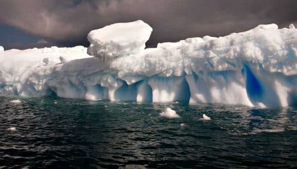 A large iceberg floating in ocean water with a blue hue and cloudy sky above. - Olive Oil Times