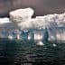 A large iceberg floating in ocean water with a blue hue and cloudy sky above. - Olive Oil Times