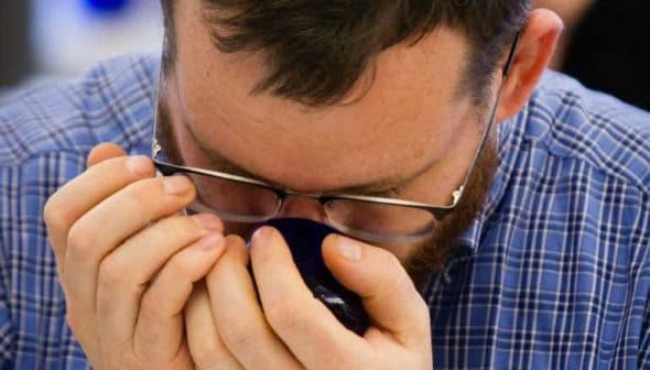 Individual with glasses holding a coffee cup close to their face, inhaling the aroma. - Olive Oil Times