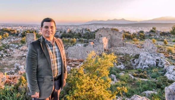 A man wearing a suit stands in front of ancient ruins during sunset with mountains in the distance. - Olive Oil Times