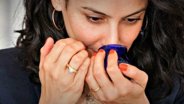 Woman with dark hair holding a blue cup close to her face, preparing to smell its contents. - Olive Oil Times