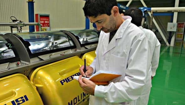 Man in a lab coat taking notes while observing yellow machinery in a facility. - Olive Oil Times