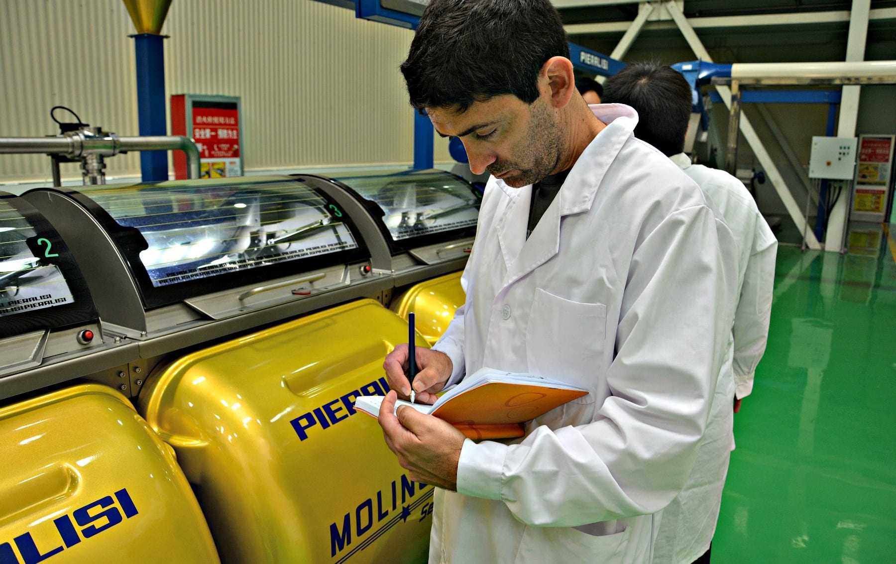 Man in a lab coat taking notes while observing yellow machinery in a facility. - Olive Oil Times
