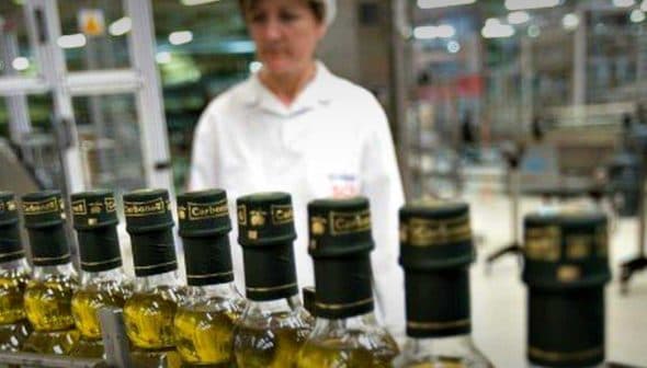 Row of olive oil bottles on a production line with a worker in the background. - Olive Oil Times