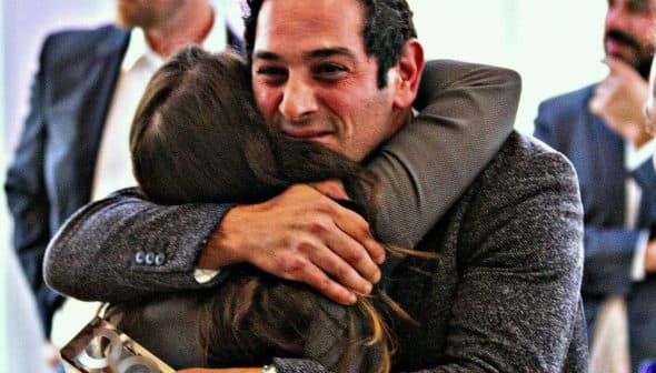 A man and a woman embracing each other in a social gathering, both smiling. - Olive Oil Times