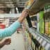 A woman reaching for a bottle of olive oil on a grocery store shelf filled with various products. - Olive Oil Times