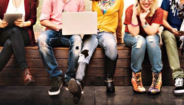 A group of young individuals sitting side by side, engaged with various devices and wearing casual clothing. - Olive Oil Times
