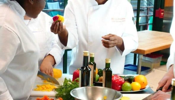 Three chefs in a culinary class preparing ingredients with fruits and vegetables on a table. - Olive Oil Times