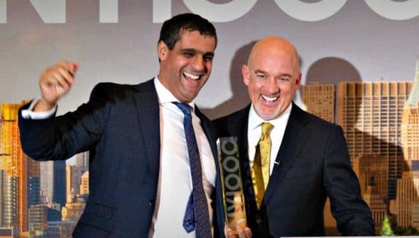 Two men smiling and celebrating on stage at an event with a city skyline backdrop. - Olive Oil Times