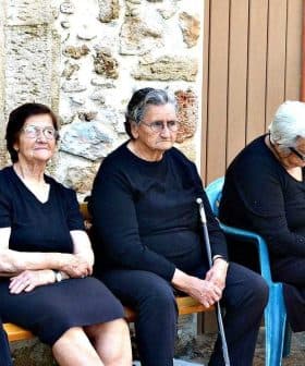 Six elderly women dressed in black and one in pink sitting on a bench outside a building. - Olive Oil Times