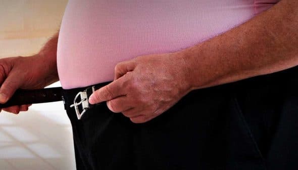 A man adjusting a black belt on his pants with both hands, wearing a pink shirt. - Olive Oil Times