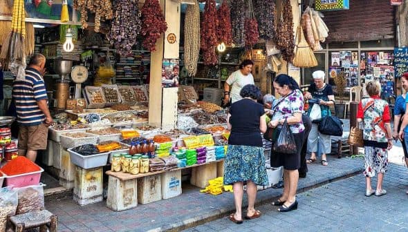 A market scene featuring various spices and dried goods displayed on tables with shoppers interacting. - Olive Oil Times