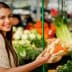 A woman smiling while holding a piece of ginger in a market filled with fresh produce. - Olive Oil Times