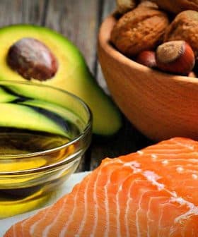 Raw salmon fillet placed on a white surface with a bowl of nuts and sliced avocado in the background. - Olive Oil Times