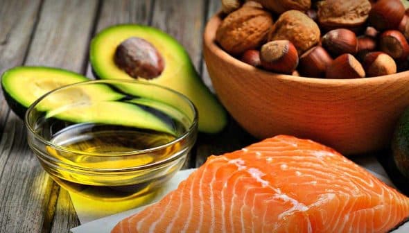 Raw salmon fillet placed on a white surface with a bowl of nuts and sliced avocado in the background. - Olive Oil Times