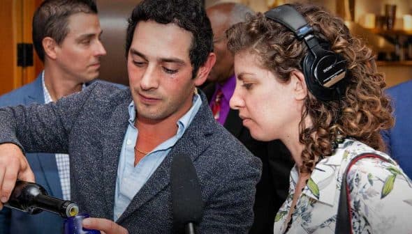 A man pouring olive oil into a glass while a woman observes at an event. - Olive Oil Times