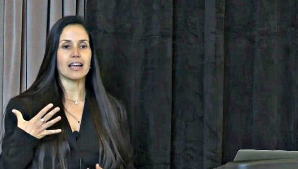 A woman with long hair speaking at a podium during a presentation at a conference. - Olive Oil Times