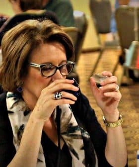 Woman holding a small cup while tasting olive oil at a dining table with flags in the background. - Olive Oil Times