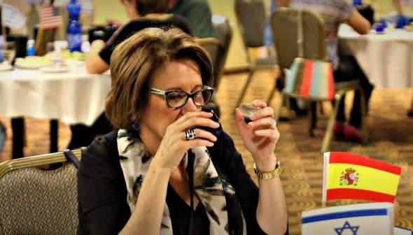 Woman holding a small cup while tasting olive oil at a dining table with flags in the background. - Olive Oil Times