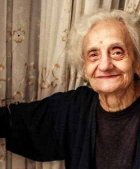 Elderly woman with gray hair smiling while standing at a doorway with curtains. - Olive Oil Times