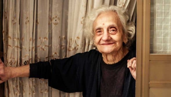 Elderly woman with gray hair smiling while standing at a doorway with curtains. - Olive Oil Times