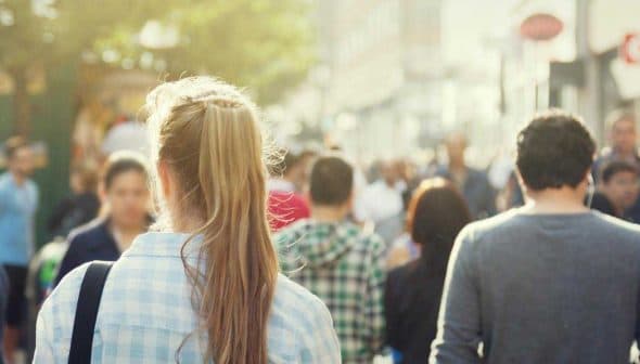 A view of a busy street with people walking in various directions, including a woman with long hair in a plaid shirt. - Olive Oil Times