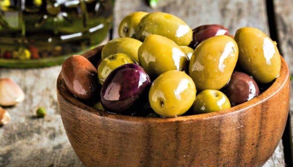 A wooden bowl filled with various types of olives including green and black varieties. - Olive Oil Times