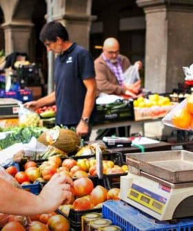 A vendor weighing apples at a market while another vendor assists a customer in the background. - Olive Oil Times
