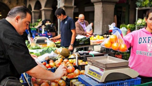 A vendor weighing apples at a market while another vendor assists a customer in the background. - Olive Oil Times
