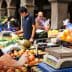 A vendor weighing apples at a market while another vendor assists a customer in the background. - Olive Oil Times