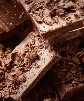 Pieces of dark chocolate stacked with chocolate shavings scattered around on a wooden surface. - Olive Oil Times