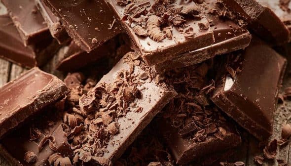 Pieces of dark chocolate stacked with chocolate shavings scattered around on a wooden surface. - Olive Oil Times