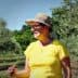 Woman wearing a yellow shirt and hat standing among olive trees in an olive grove. - Olive Oil Times