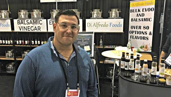 Man standing in front of a booth displaying balsamic vinegar and olive oil products at a trade show. - Olive Oil Times