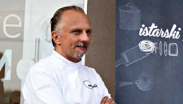 Chef wearing a white uniform standing in front of a sign for a culinary festival. - Olive Oil Times
