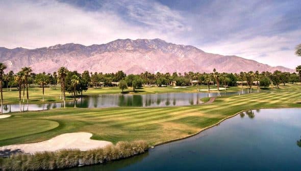A panoramic view of a golf course featuring water hazards and palm trees with mountains in the background. - Olive Oil Times