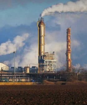 Industrial complex featuring multiple smoke stacks emitting white smoke against a blue sky. - Olive Oil Times