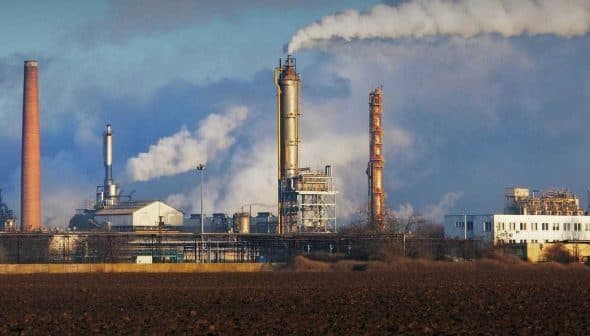 Industrial complex featuring multiple smoke stacks emitting white smoke against a blue sky. - Olive Oil Times