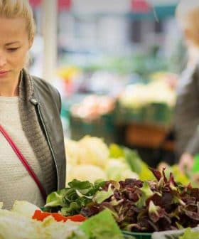 Woman examining fresh vegetables at a market stall surrounded by other shoppers. - Olive Oil Times