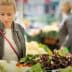 Woman examining fresh vegetables at a market stall surrounded by other shoppers. - Olive Oil Times