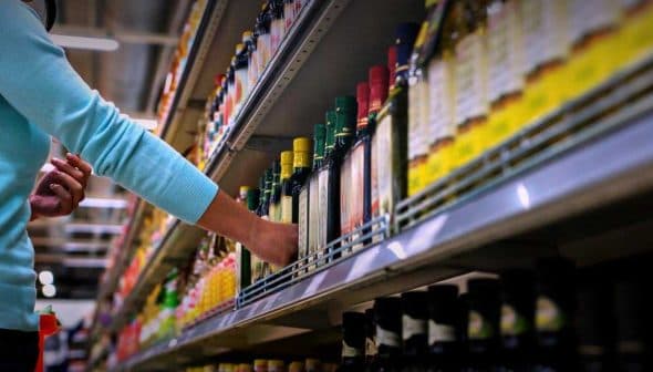 Individual reaching for a bottle of olive oil on a grocery store shelf with various products. - Olive Oil Times