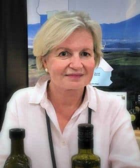 A woman with short blonde hair sitting at a table with olive oil bottles at an exhibition. - Olive Oil Times