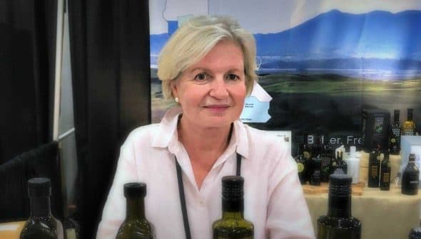 A woman with short blonde hair sitting at a table with olive oil bottles at an exhibition. - Olive Oil Times