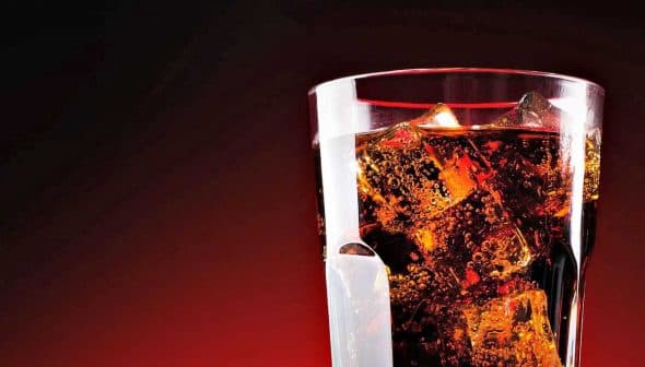 A glass filled with cola and ice cubes against a dark background. - Olive Oil Times