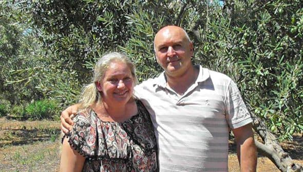 A couple standing together in an olive grove, smiling at the camera. - Olive Oil Times
