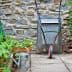 A metal wheelbarrow positioned on a stone pathway surrounded by various potted plants and a watering can. - Olive Oil Times