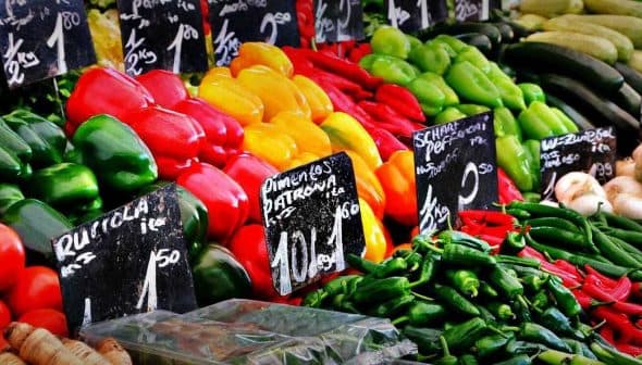 A variety of fresh vegetables including peppers, tomatoes, and cucumbers arranged at a market stall. - Olive Oil Times