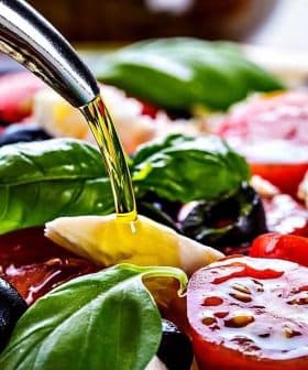 Fresh salad with tomatoes, mozzarella, olives, and basil being drizzled with olive oil. - Olive Oil Times